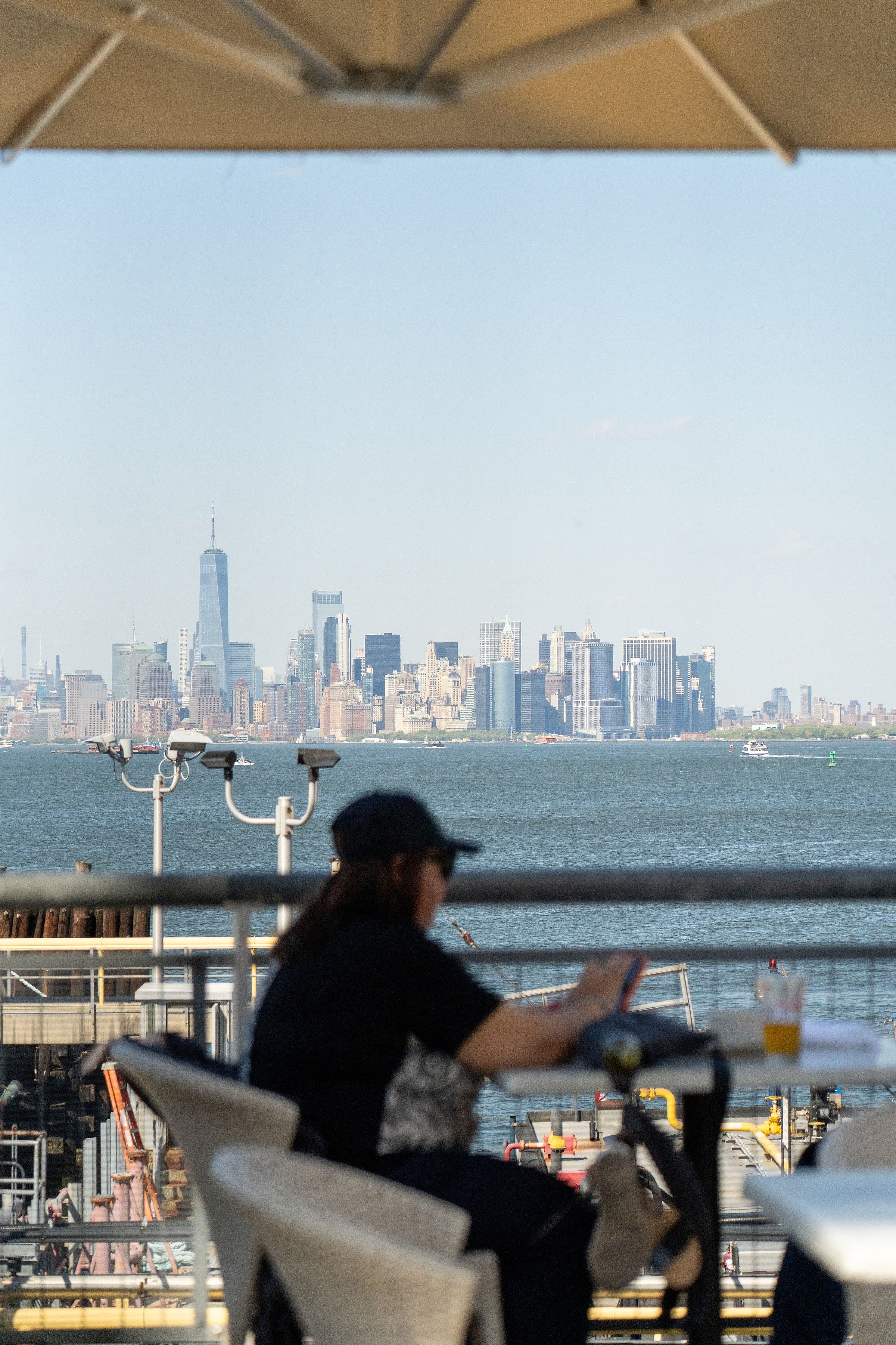 A diner under an umbrella on the Staten Island waterfront, the Manhattan skyline across the harbor